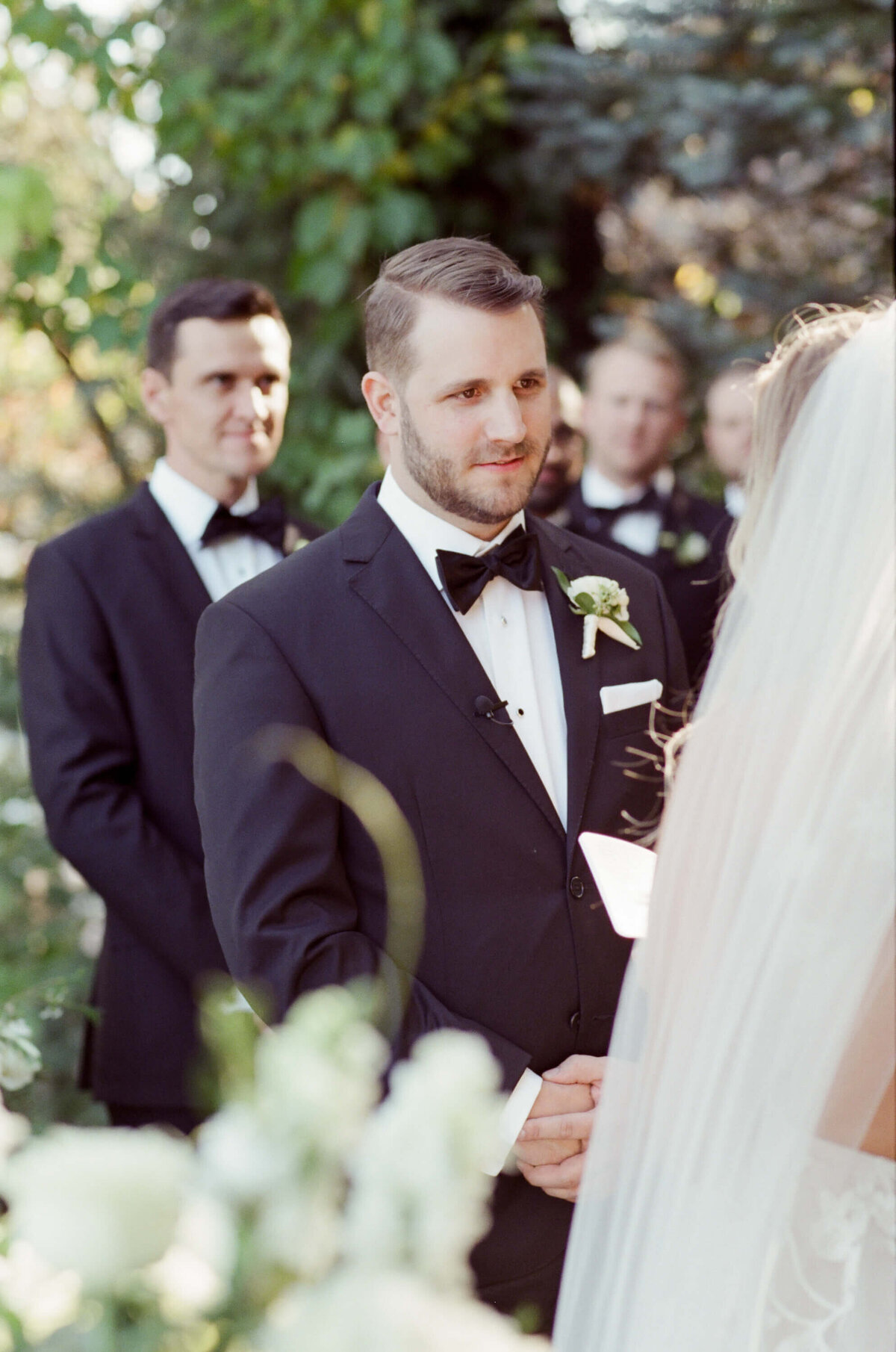 Bridegroom in elegant black suit and bow tie looks at his spouse while his best men attend the outdoor vineyard wedding.