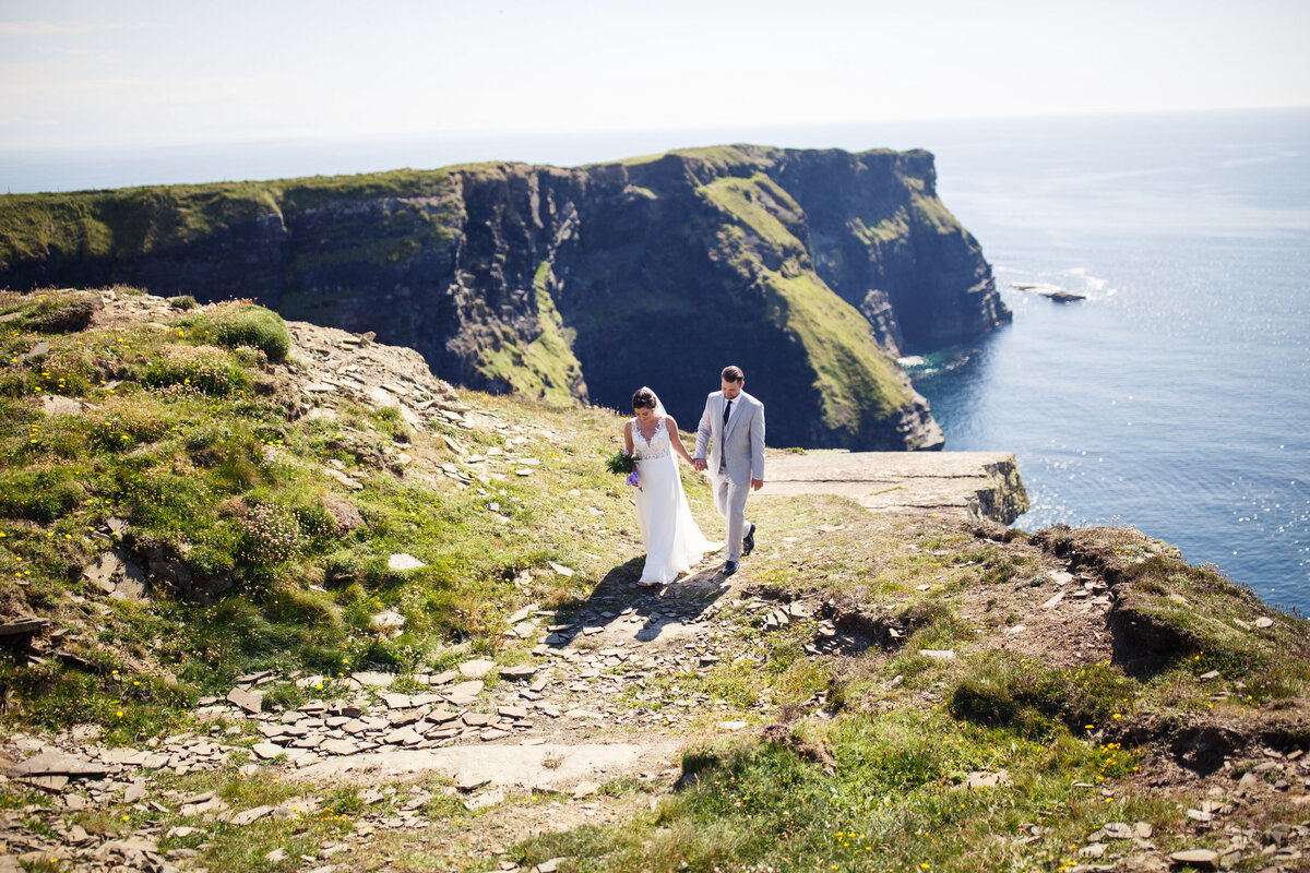 bride and groom holding hands and walking with the Cliffs of Moher behind them
