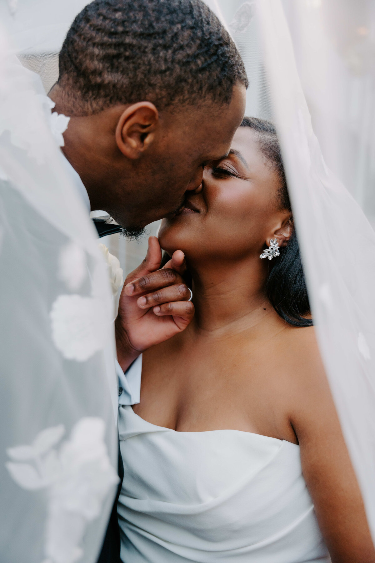 A bride and groom share a tender kiss under a delicate, flower-adorned veil. The moment is intimate and romantic, conveying love and warmth.