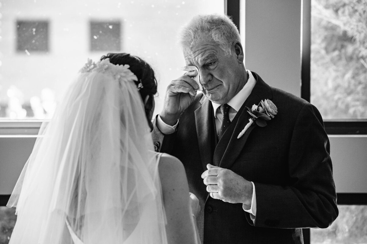 Black-and-white-image-of-a-father-wiping-tears-from-his-eyes-seeing-his-daughter-for-the-first-time-during-their-first-look-at-St-Gabriel-Catholic-Church-in-Charlotte