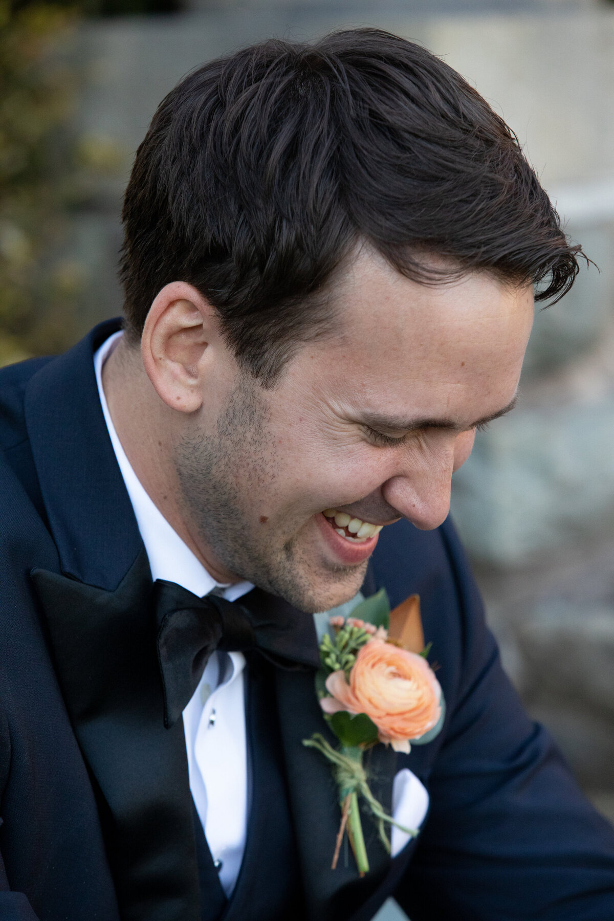 A man in a blue suit wearing an orange flower on his lapel looks down and smiles Paramount Country club wedding photographer Milestone Images