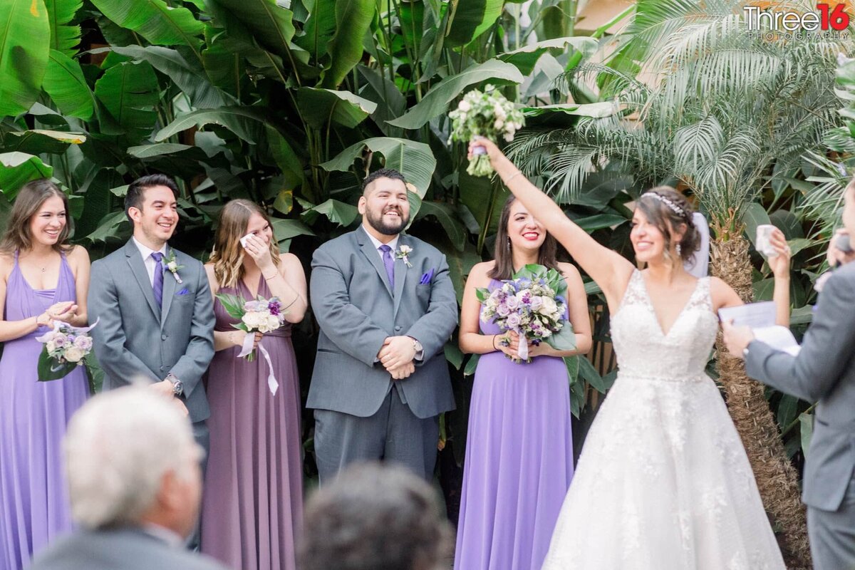 Newly married Bride lifts her arm in the air with bouquet in hand as the wedding party smiles