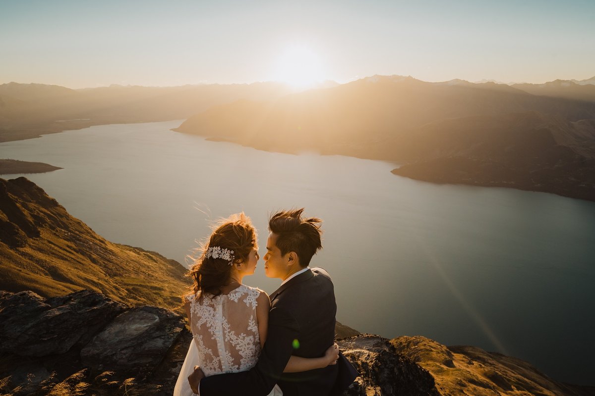 bride and groom sat on a cliff at sunset over the water