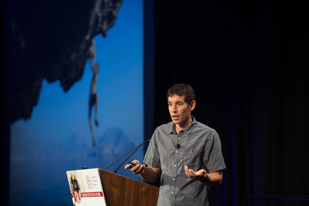 Alex Hannold speaks at a conference as a photo of him hanging from a rock is projected behind him