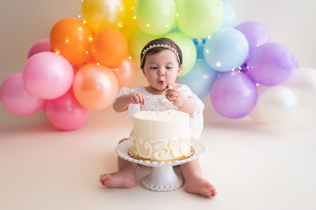 A one-year-old girl digs into a birthday cake in front of a rainbow balloon arch in our Waukesha photo studio.
