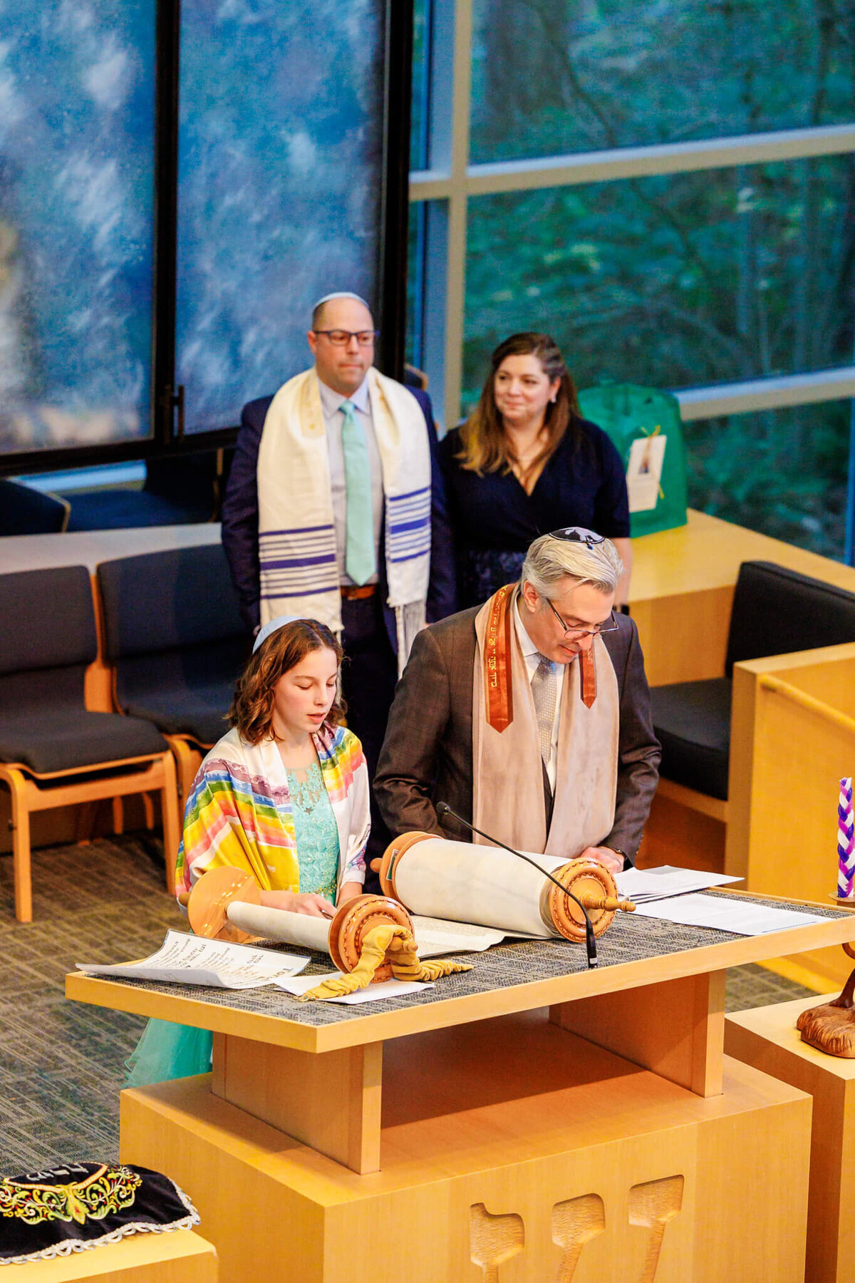 A view at a teenage girl standing at the bimah reading the torah with a rabbi