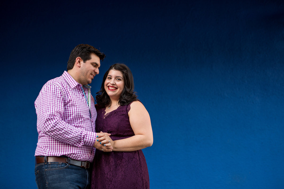 Couple on their engagement session standing in front of a blue wall and holding hands and posing for Expose The Heart Photography.