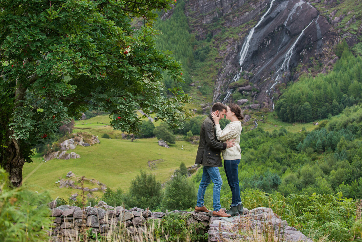 Young couple standing on an old, stone wall kissing in front of a waterfall surrounded by trees