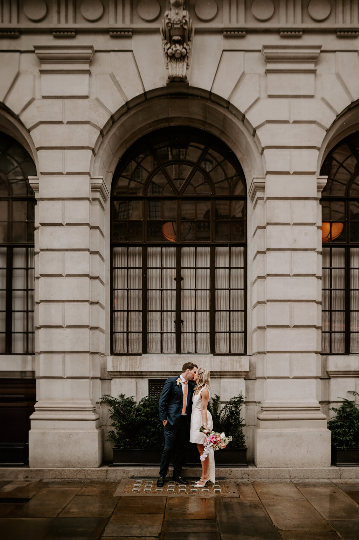 A wedding couple kiss outside The Ned in London. The Bride is wearing a short wedding dress and the groom is wearing a skinny fit navy suit.