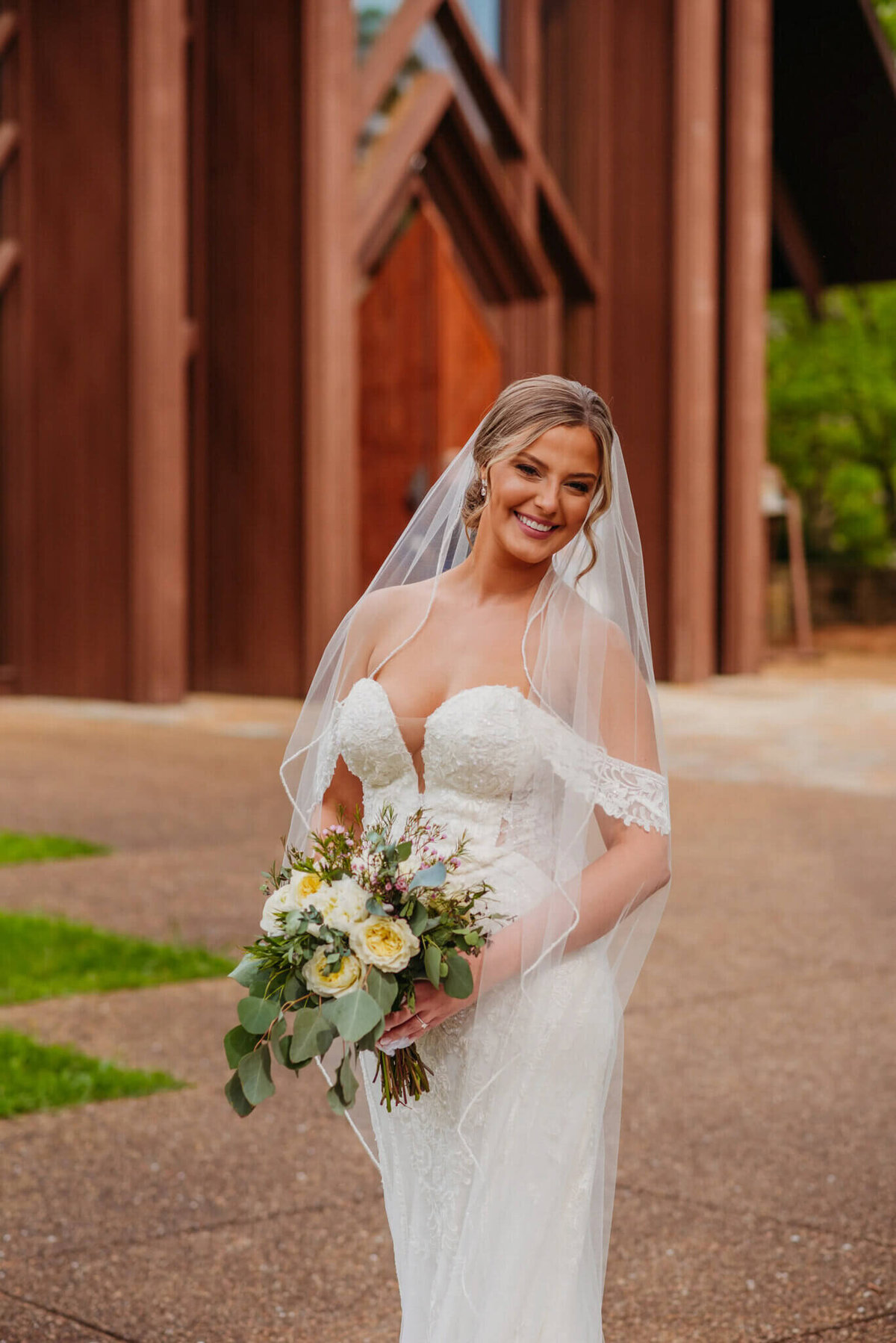 photo of a bride holding a bouquet and wearing a wedding veil while standing in front of a wooden chapel
