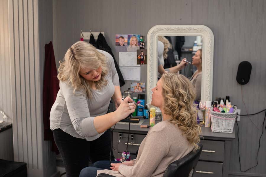 A makeup artist applies makeup to a seated woman in a salon, both reflected in a large mirror surrounded by makeup products.