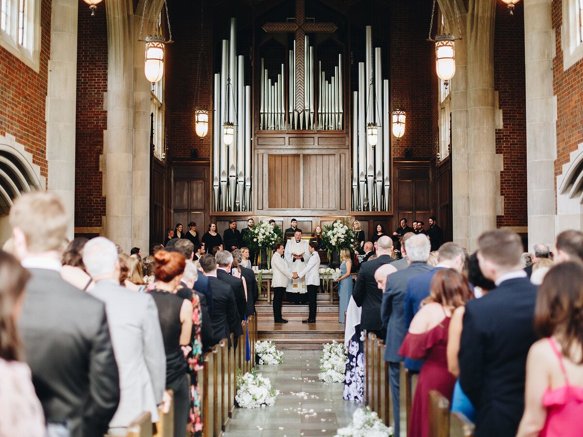 Two grooms wearing white tuxedo jackets and black pants hold hands at Scarritt Bennett in front of a large church pipe organ as wedding guests look on. The aisle is lined with round white floral arrangements and the altar has tall white floral arrangements.