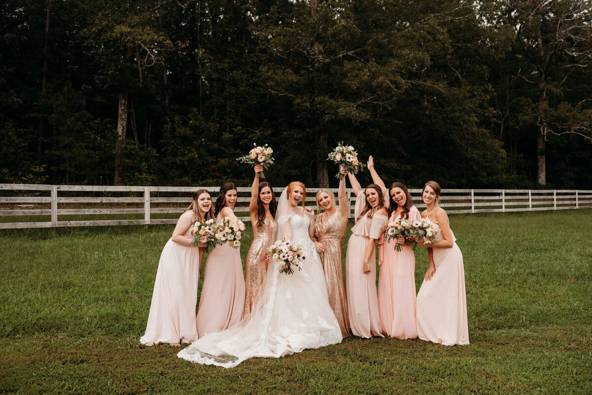 Photo of a bride and bridesmaids wearing pink dresses cheering with a wooden fence in the background