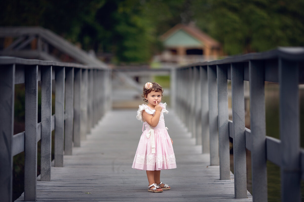 A young toddler girl in a pink dress with bows places a finger in front of her mouth while walking on a bridge in a park