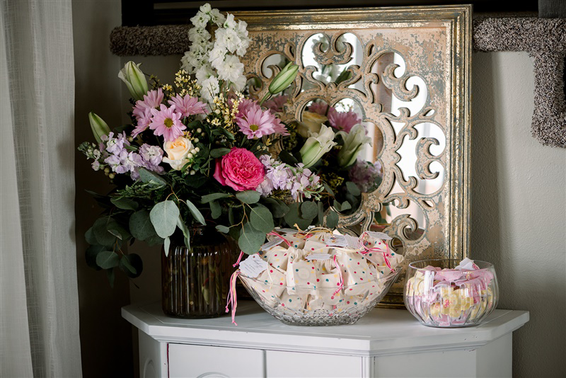 Pink and yellow floral centerpiece arranged against a mirror with elegant favors in a crystal bowl beneath it, displayed on a white cabinet at a girl’s baby shower.