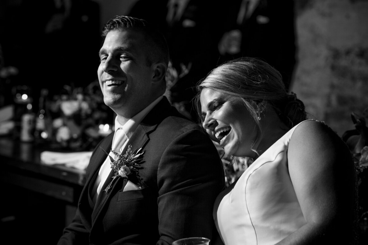 Black-and-white-image-of-a-bride-and-groom-laughing-during-reception-toasts-at-The-Providence-Cotton-Mill