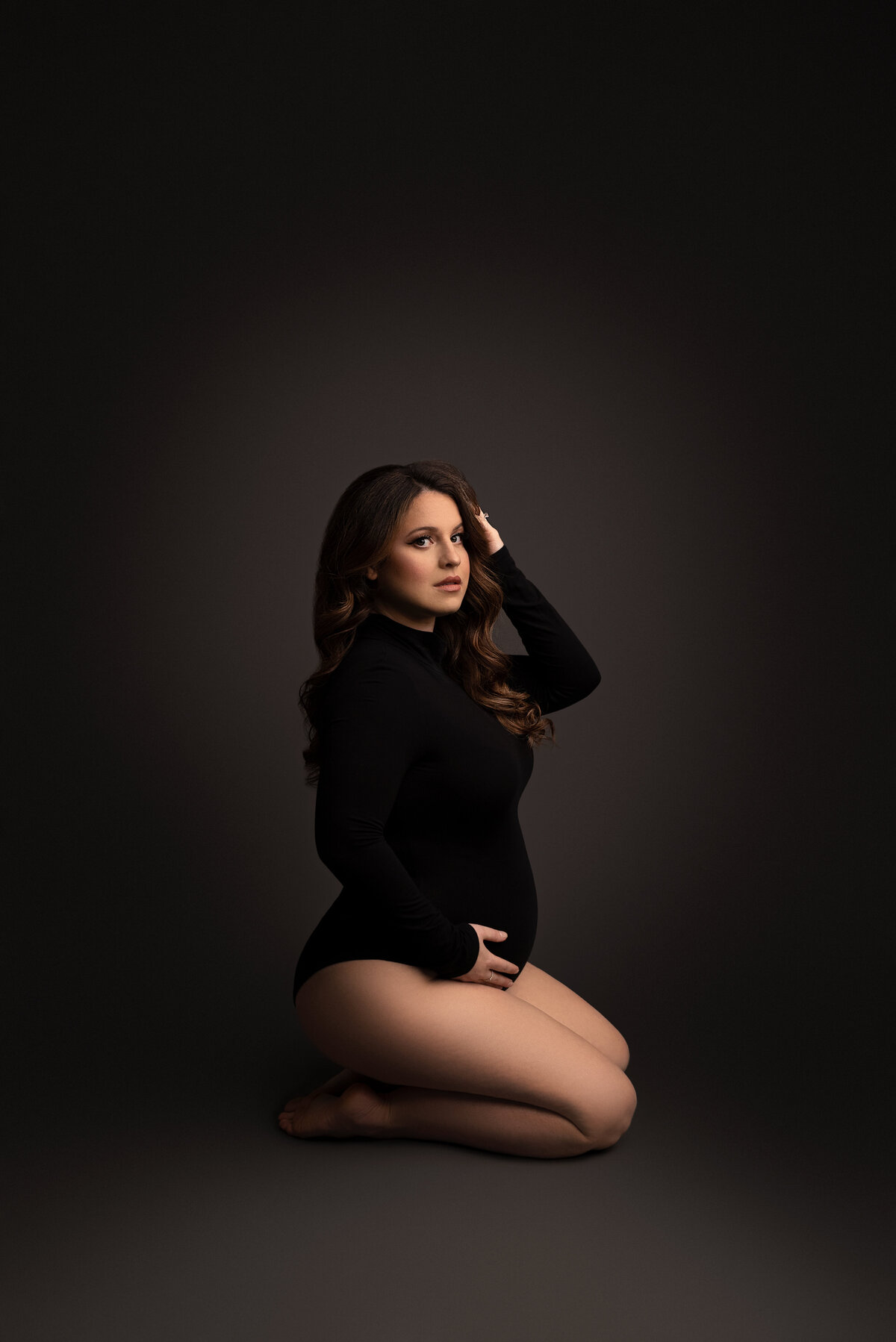 Maternity photo by Katie Marshall, Philadelphia Main Line's premier maternity photographer. Woman in a long-sleeve black bodysuit with bare legs, sitting on her knees, angled away from the camera. Her forearm supports her baby bump, while her back hand brushes hair off her face. Her head is gently tilted toward the camera, set against a grey backdrop, creating an intimate and artistic composition.