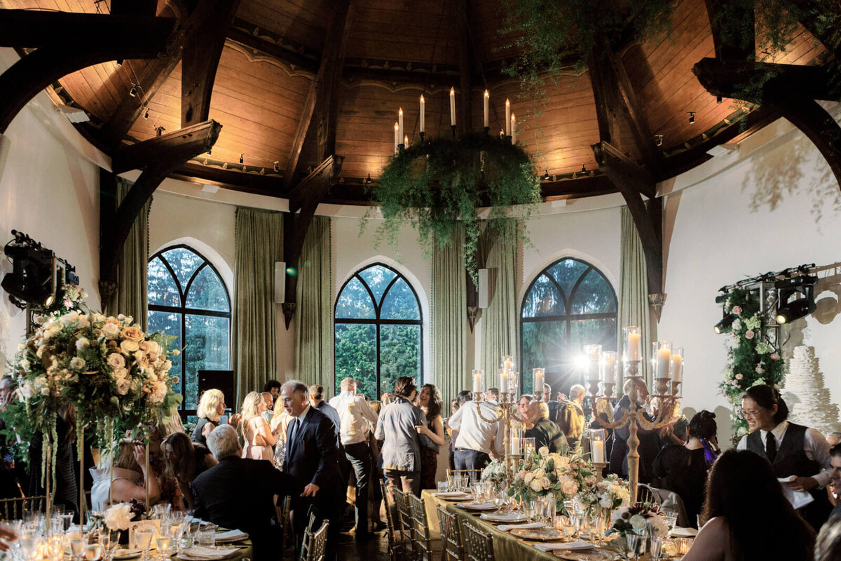 Wedding guests are chatting inside an elegant wedding reception room, with a candle chandelier and over-sized windows.
