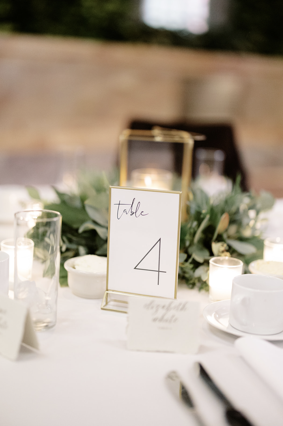 Gold and white decorations accompanied by minimalist table numbers and name cards are paired with lanterns and glass tea lights for elegant wedding reception centerpieces.