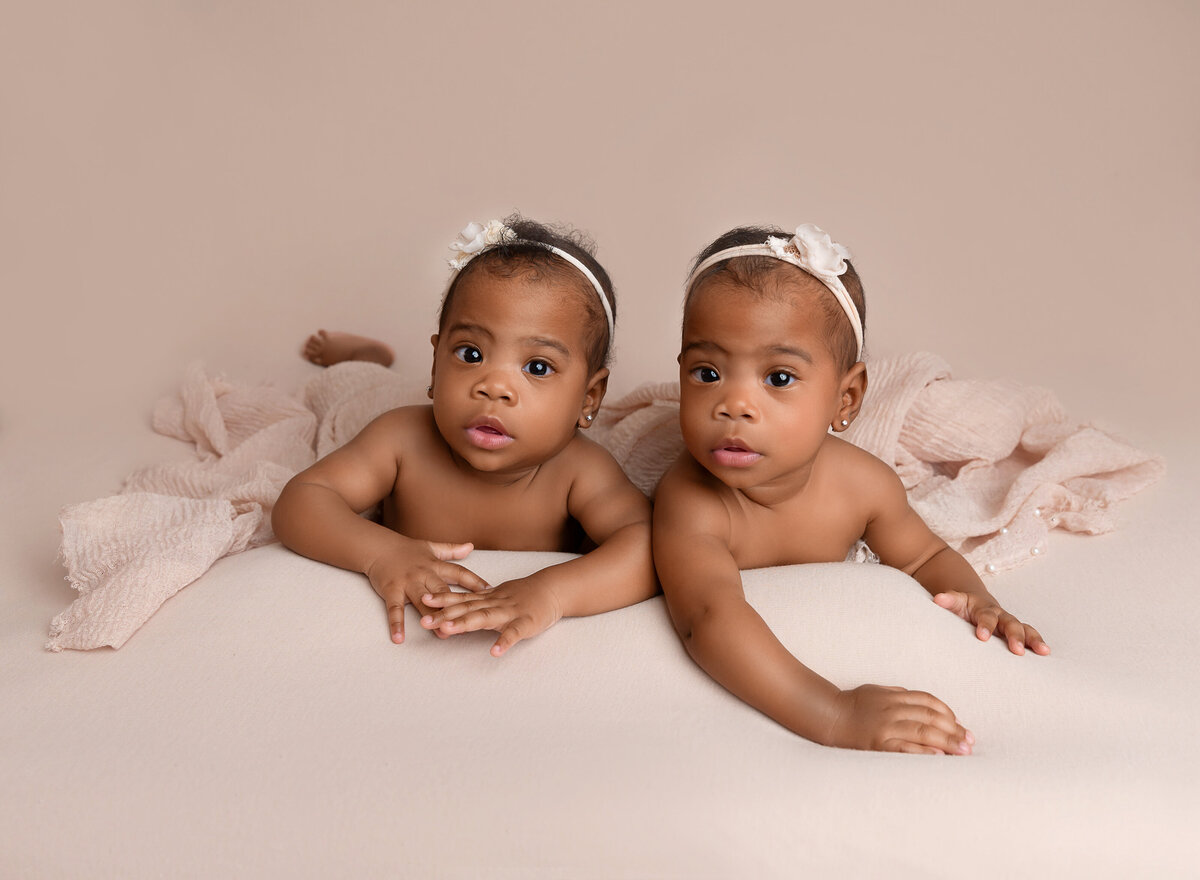 6-month old twin girls pose for a baby milestone photoshoot. Babies are propping their heads up and looking at the camera with a serious expression.