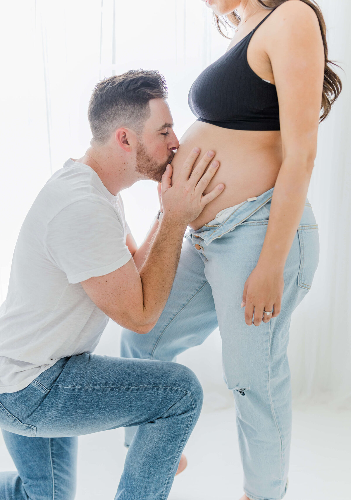 Couple standing together while man kneels down and kisses pregnant wife's stomach