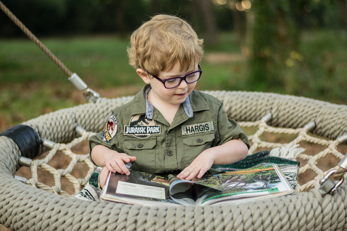 A young boy reads a dinosaur book in a large swing.