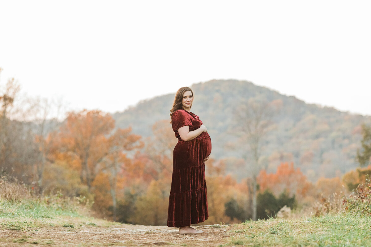 pregnant mom in red velvet dress stands in front of a mountain in fall with with orange leaves