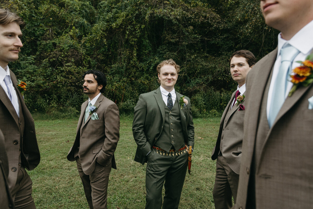 Groomsmen standing together in earthy-toned suits at an outdoor fall wedding at The Nest in Northwest Arkansas, featuring the groom in an olive green suit and natural boutonnières designed by Fleurish NWA wedding florist.