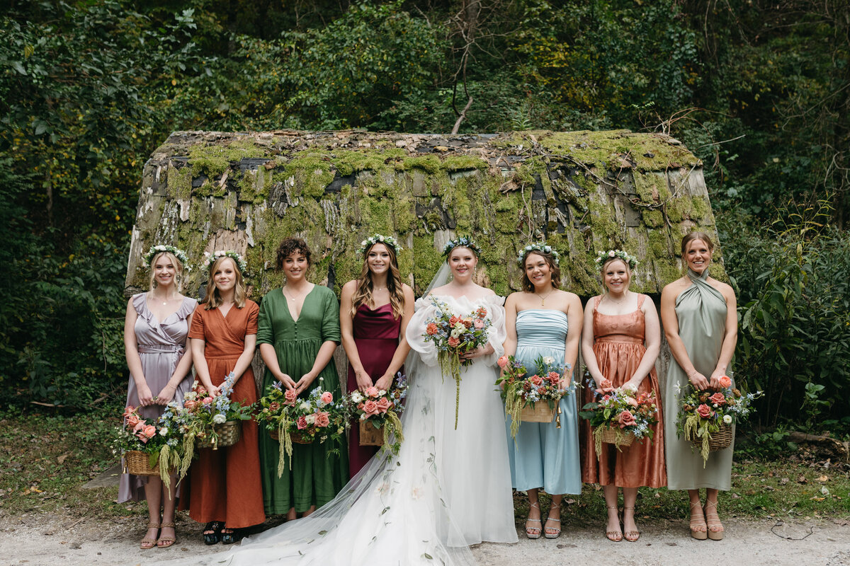 Bridesmaids in mixed earthy-tone dresses—sage, rust, burgundy, and blue—standing with the bride in front of a moss-covered structure, each holding overflowing wildflower-inspired bouquets arranged by a wedding florist in Arkansas.