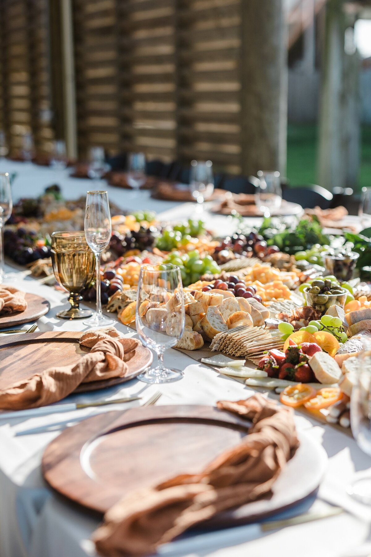 A detail shot of a reception table at a wedding in Crystal Beach, Texas. Multiple place settings surround the edges of the table using wooden plates, napkins, cutlery, and glasses. The middle of the table is made up of a large assortment of meats, cheeses, crackers, and fruits all in one large spread.