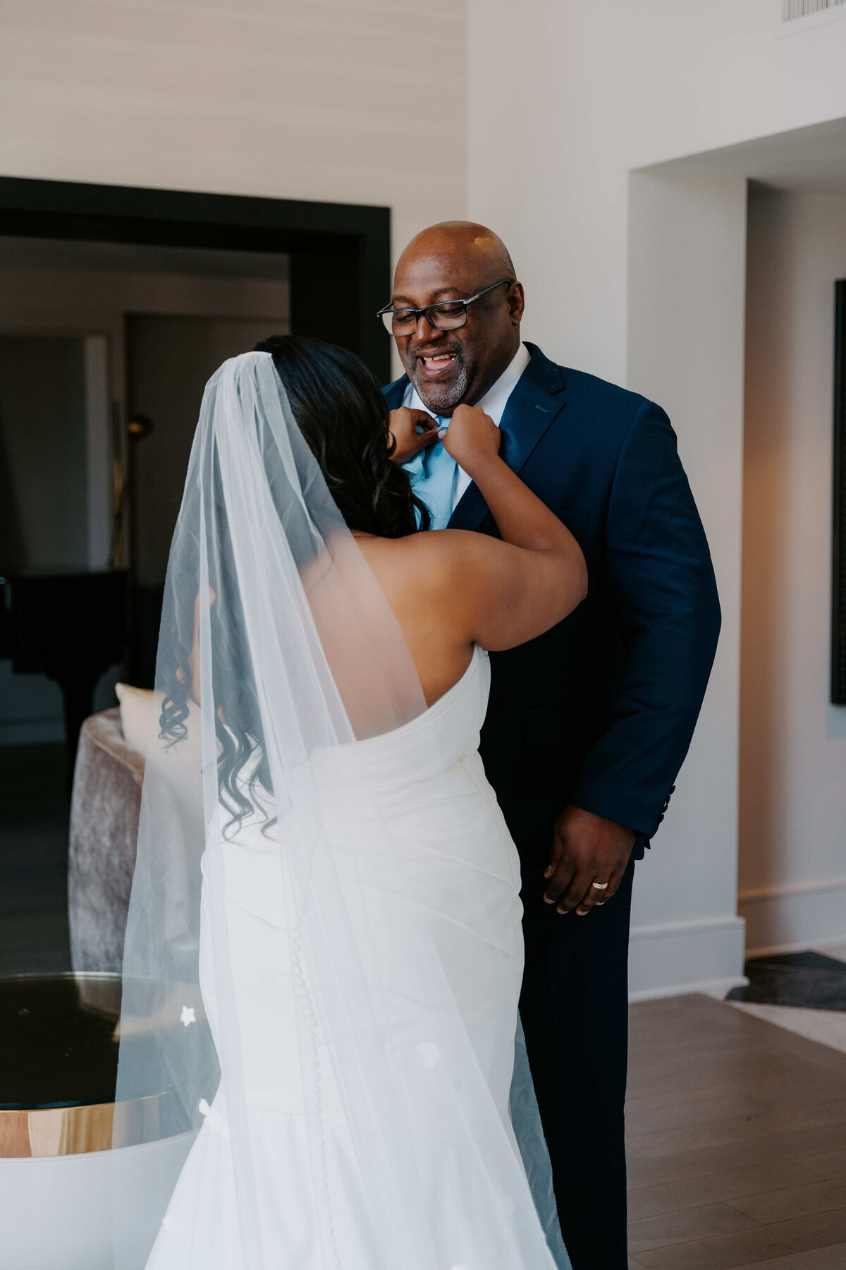 A bride in a white dress and veil adjusts a man's blue tie in a warmly lit room. Both are smiling, conveying joy and warmth.