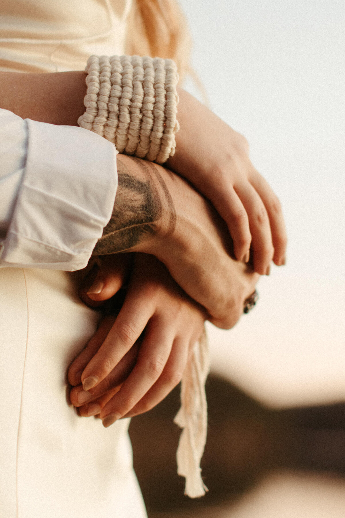 Close up view of a bride with a boho bracelet holding her groom's hands at sunset