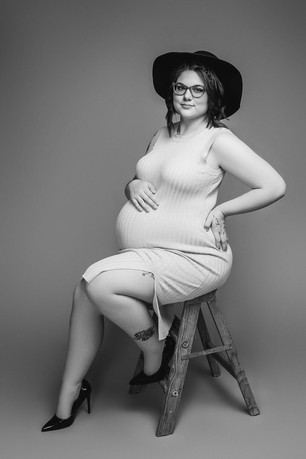 A pregnant woman in a bodycon dress and a black floppy hat sits on a wooden ladder in front of a neutral-toned tan backdrop for her glamour maternity photoshoot, captured by a photographer in Bemidji, Minnesota