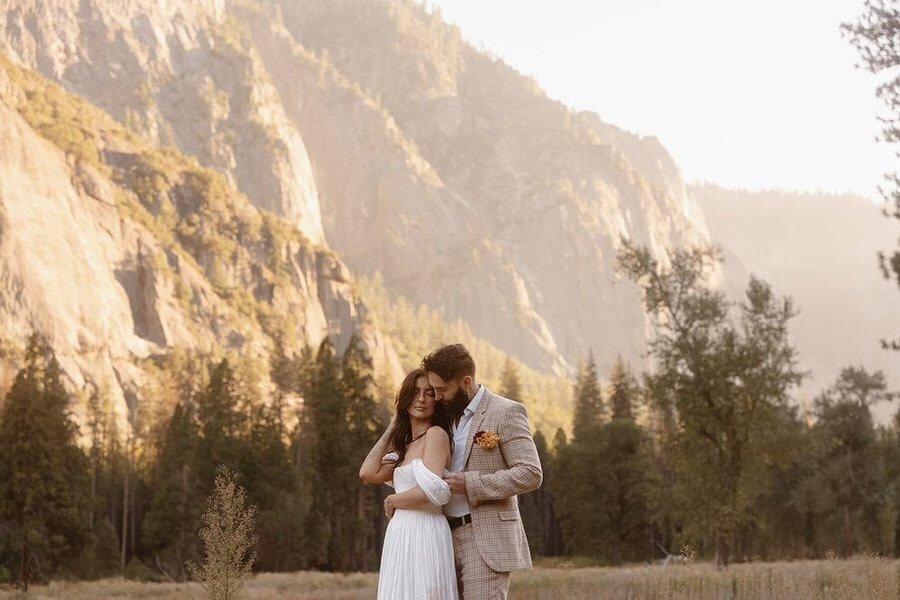 A couple stands nestled in each other’s arms, with cliffs and pine trees behind them in warm evening light.