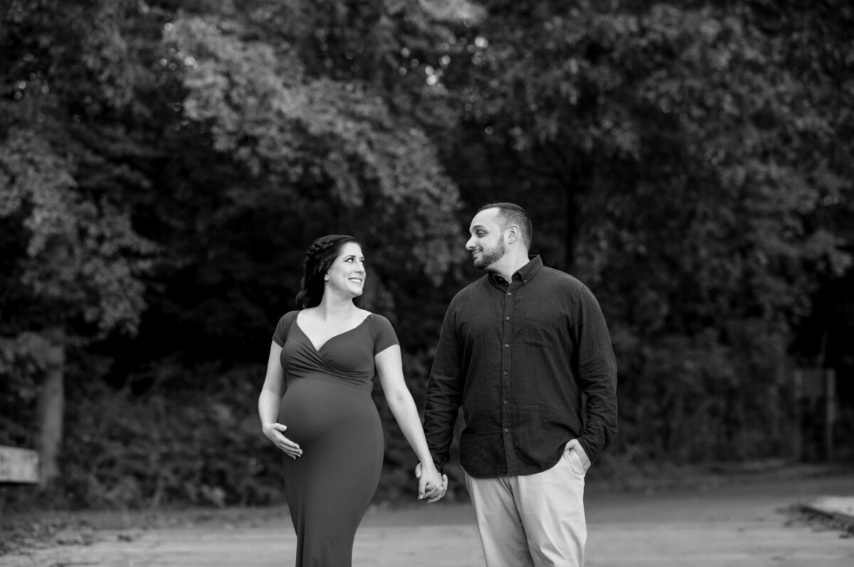 Man and woman post for a maternity photo, looking at each other in front of trees