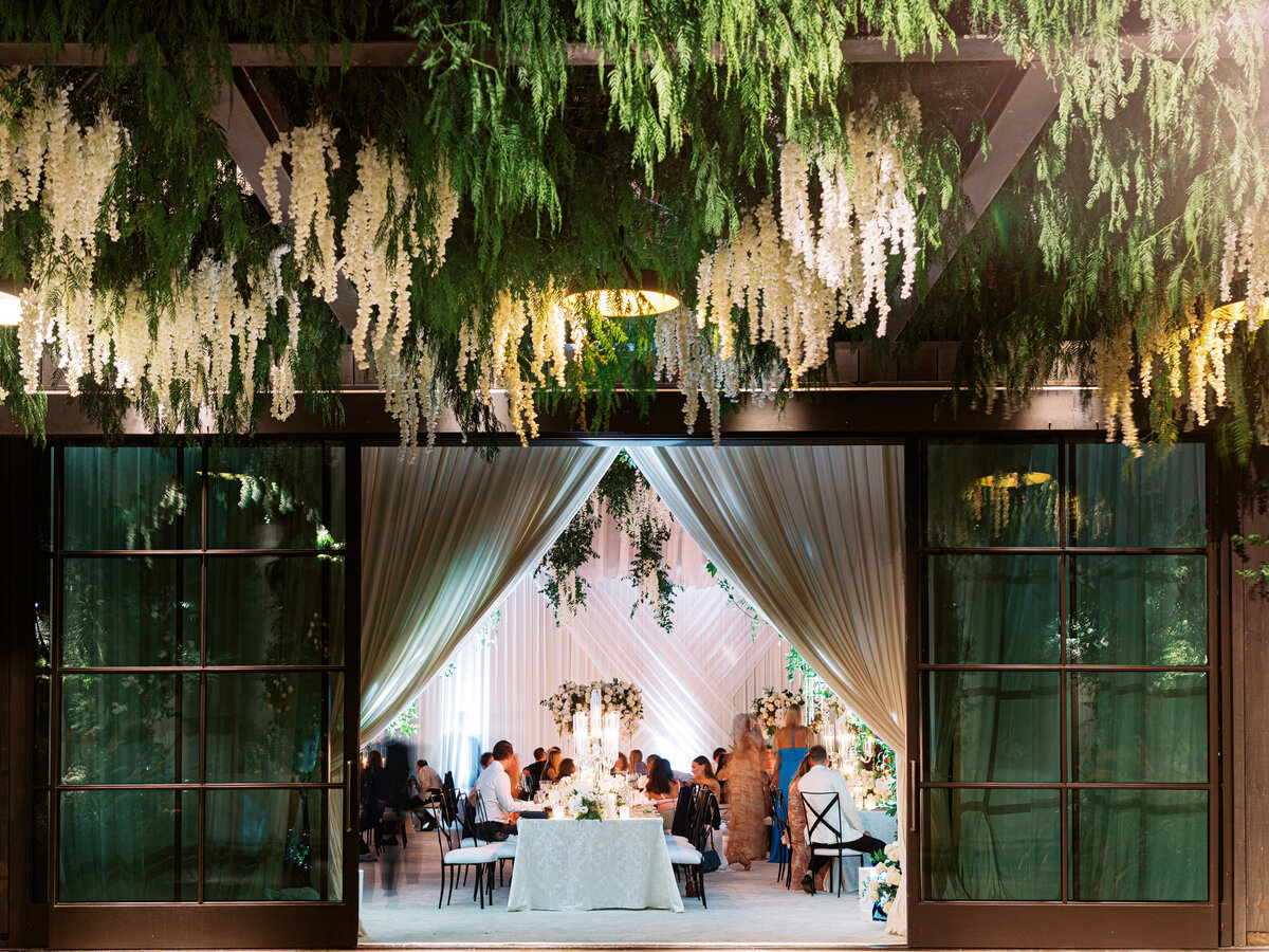 Luxurious indoor, outdoor wedding reception in Ojai Valley California with beautiful greenery among the ceiling and white drapery.