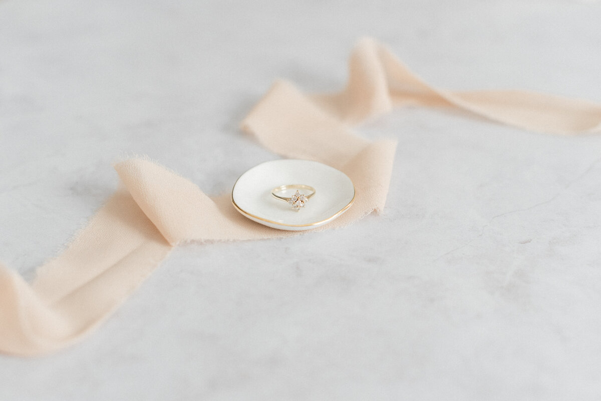 engagement ring sitting in small ivory dish on top of pale pink silk ribbon on a light marble backdrop