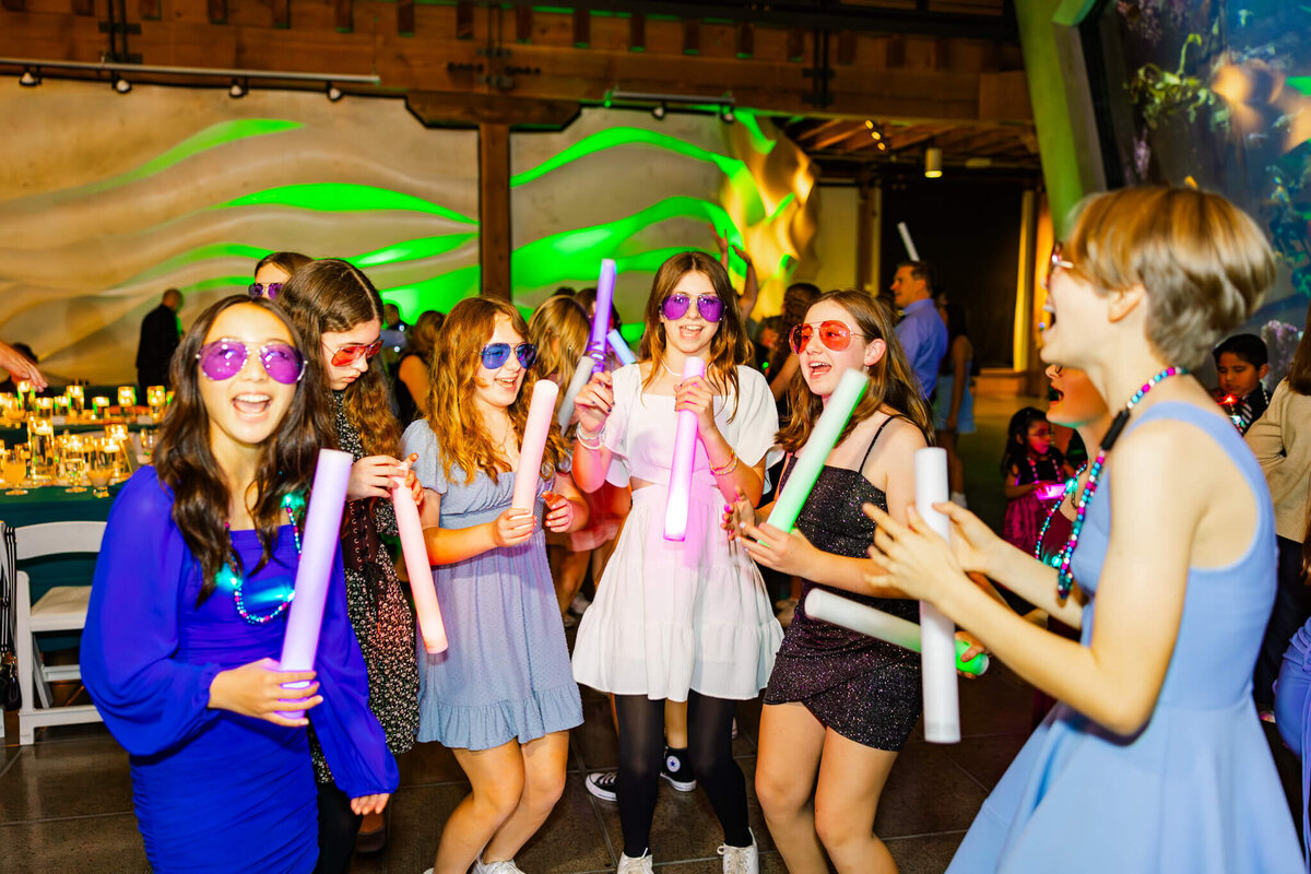 A group of teen girls dance and sing in colorful sunglasses and glow tubes