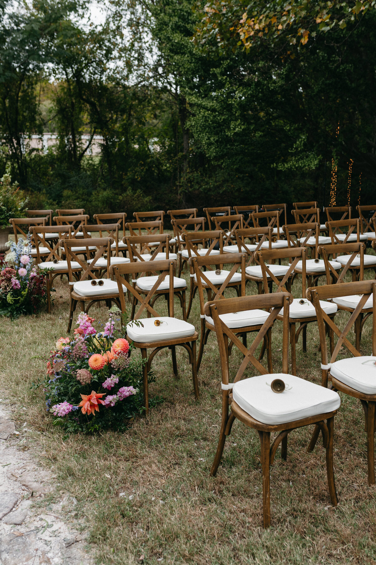 Rows of wooden cross-back ceremony chairs set with petite handbells and surrounded by colorful garden-style aisle meadow florals for an outdoor wedding at The Nest in Northwest Arkansas.