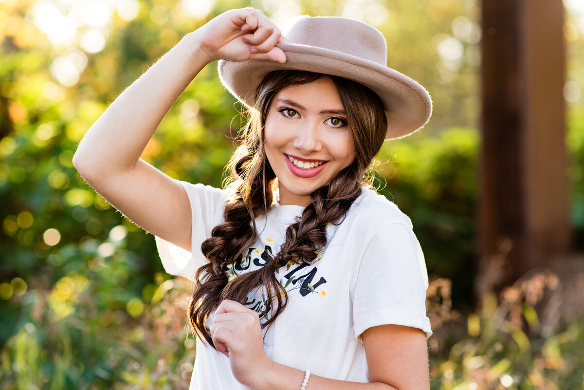 Richmond senior girl with braids and tan hat poses in field of tall grass at sunset.