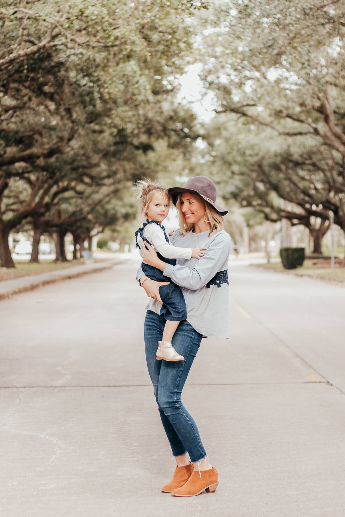 Stylish mom holds her toddler daughter in a beautiful street in Friendswood, Texas wearing a hat and  booties while daughter has messy pigtails and overalls.