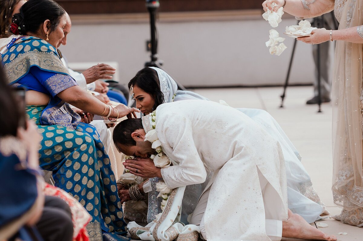 The Hindu bride and groom kneel at the feet of their elders as they receive blessings. The groom is wearing a white sherwani with a white floral varmala garland. The bride is wearing a light blue dupatta veil. The mother of the bride, wearing a turquoise and royal blue saree with gold accents places her hands on their heads to bless them as the officiant wearing a gold lehenga choli sprinkles them with white flower petals.