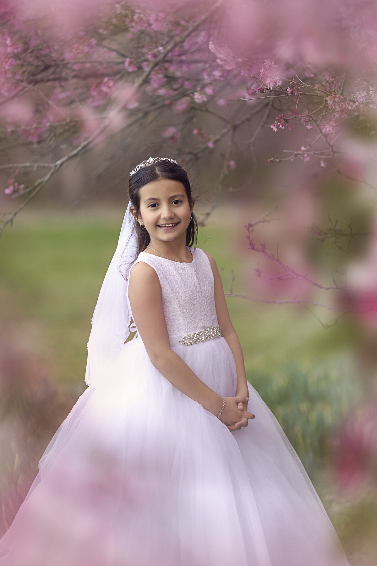 A young girl in a long pink dress stands under some pink tree blossoms smilingNew Jersey Communion Portrait Photographer