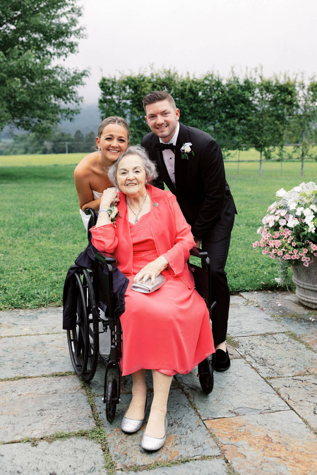 The bride and the groom are with a happy elderly woman in a wheelchair on the beautiful grounds of Lion Rock Farm, CT.