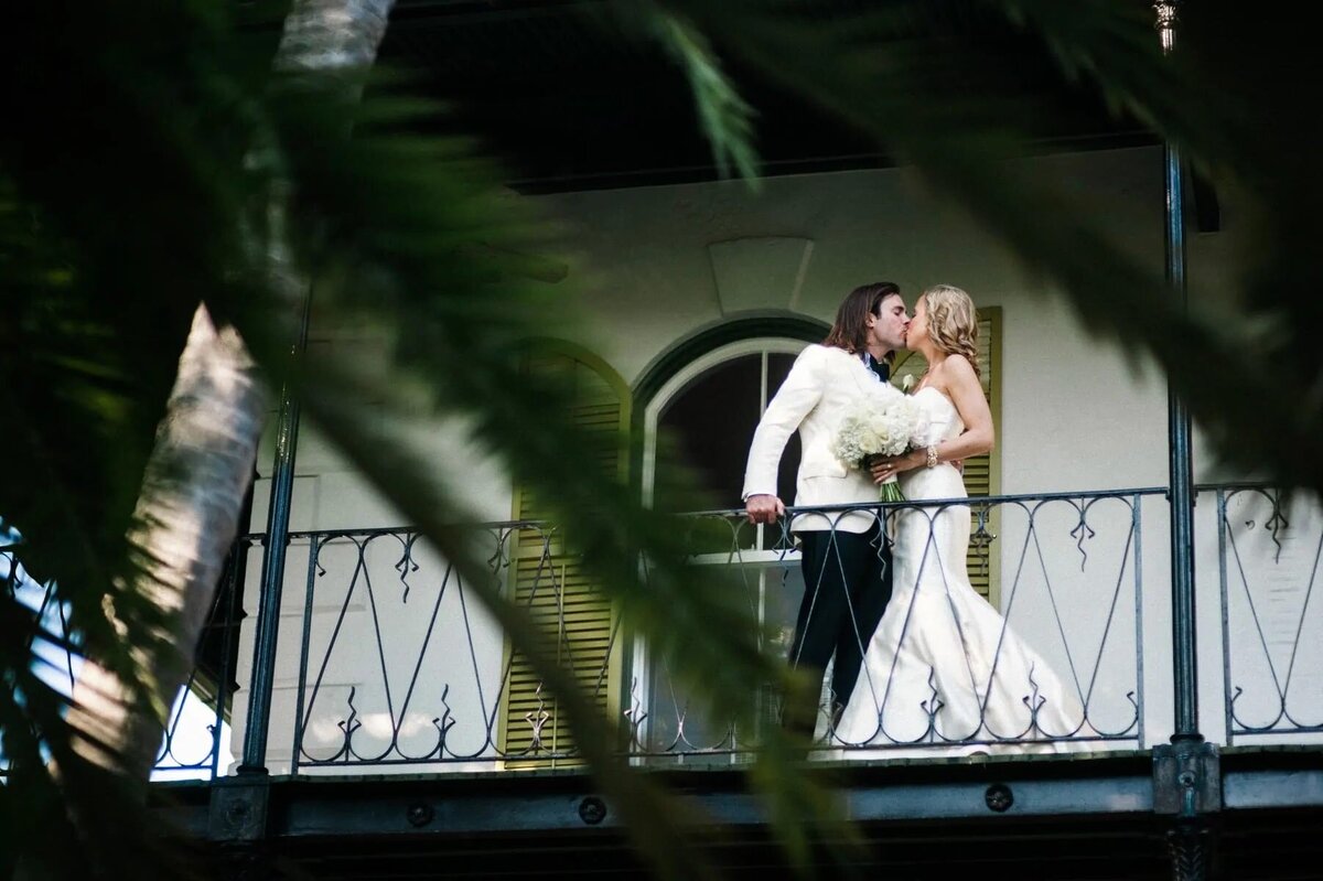 Hidden behind tropical foliage, a bride and groom stand on a balcony sharing an intimate kiss, framed by the vibrant green leaves.