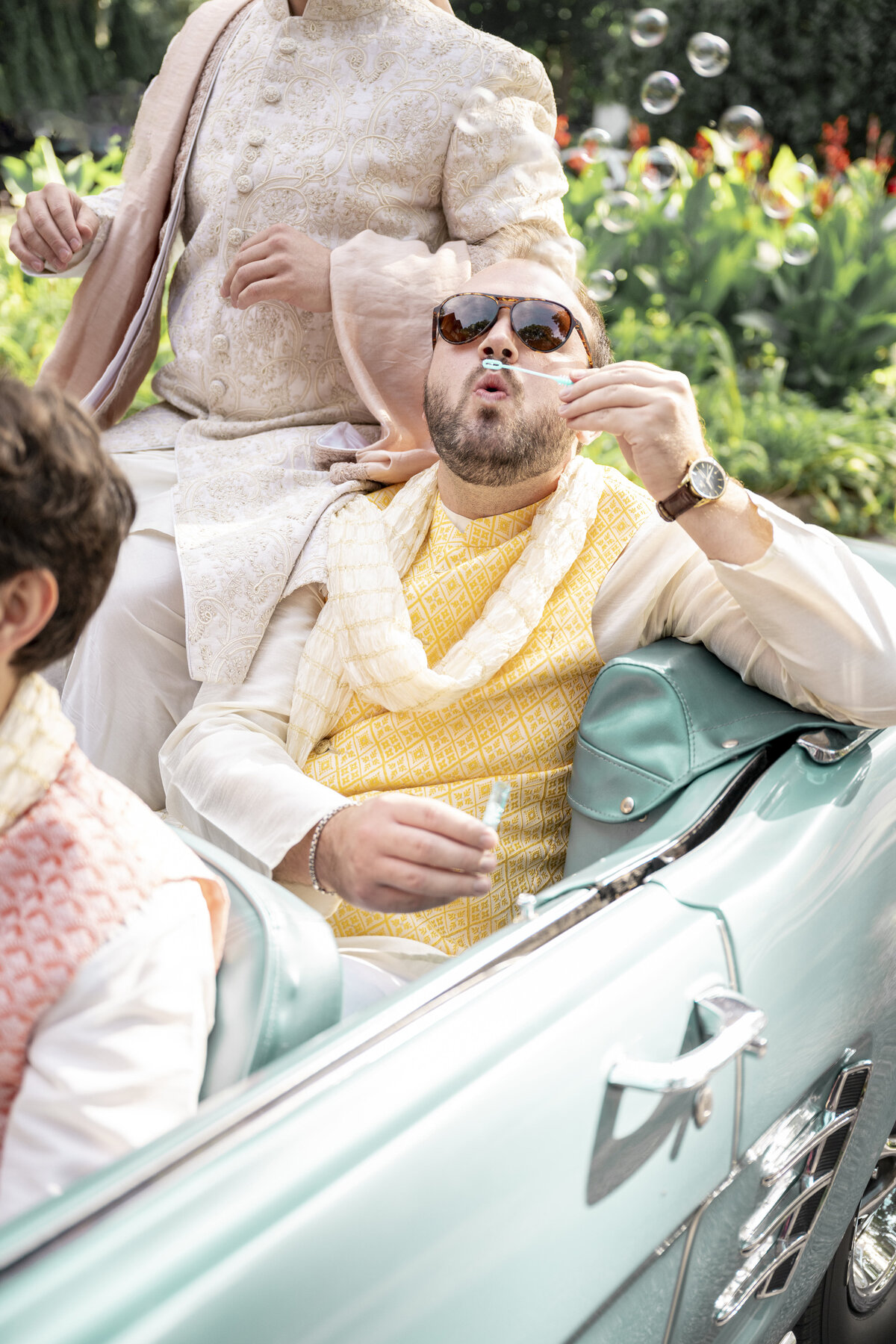 A man wearing sunglasses and traditional South Asian clothing blows bubbles while sitting in a teal convertible. He is surrounded by greenery and other people also dressed in similar attire.