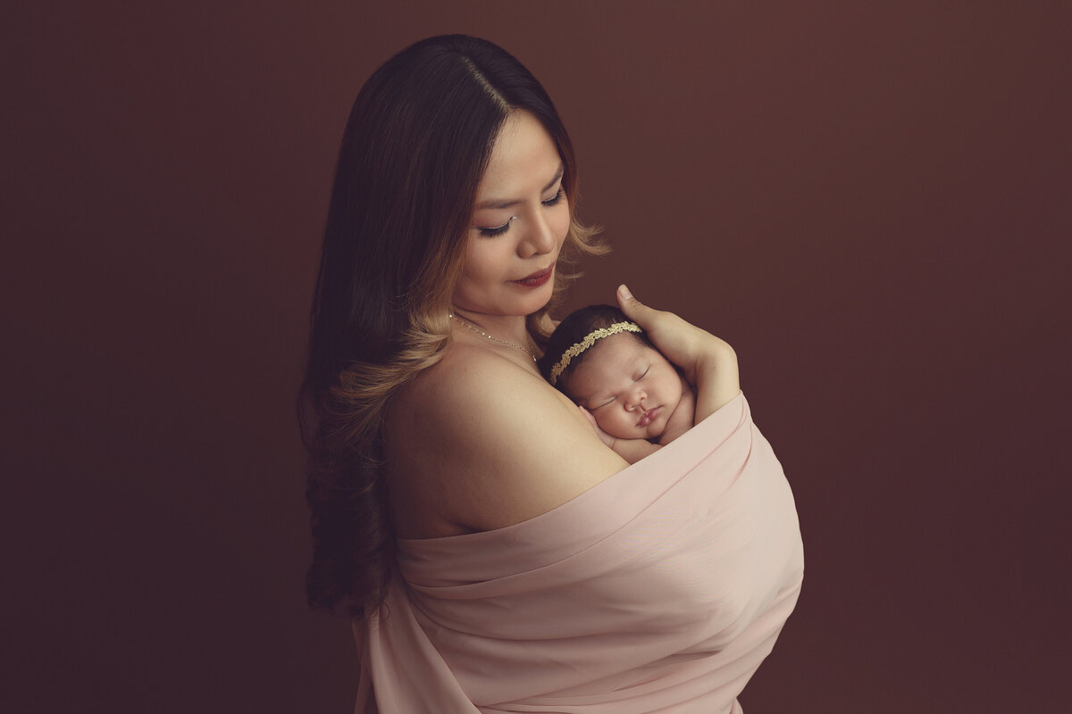 A mother in a pink dress cradles her sleeping newborn daughter against her chest while standing in a studio