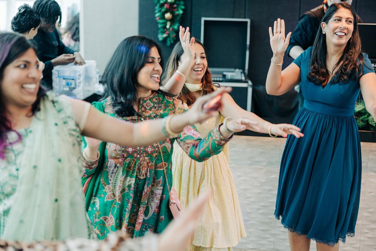 A group of women joyfully dancing together at a celebration, one in a teal dress leading with a smile.