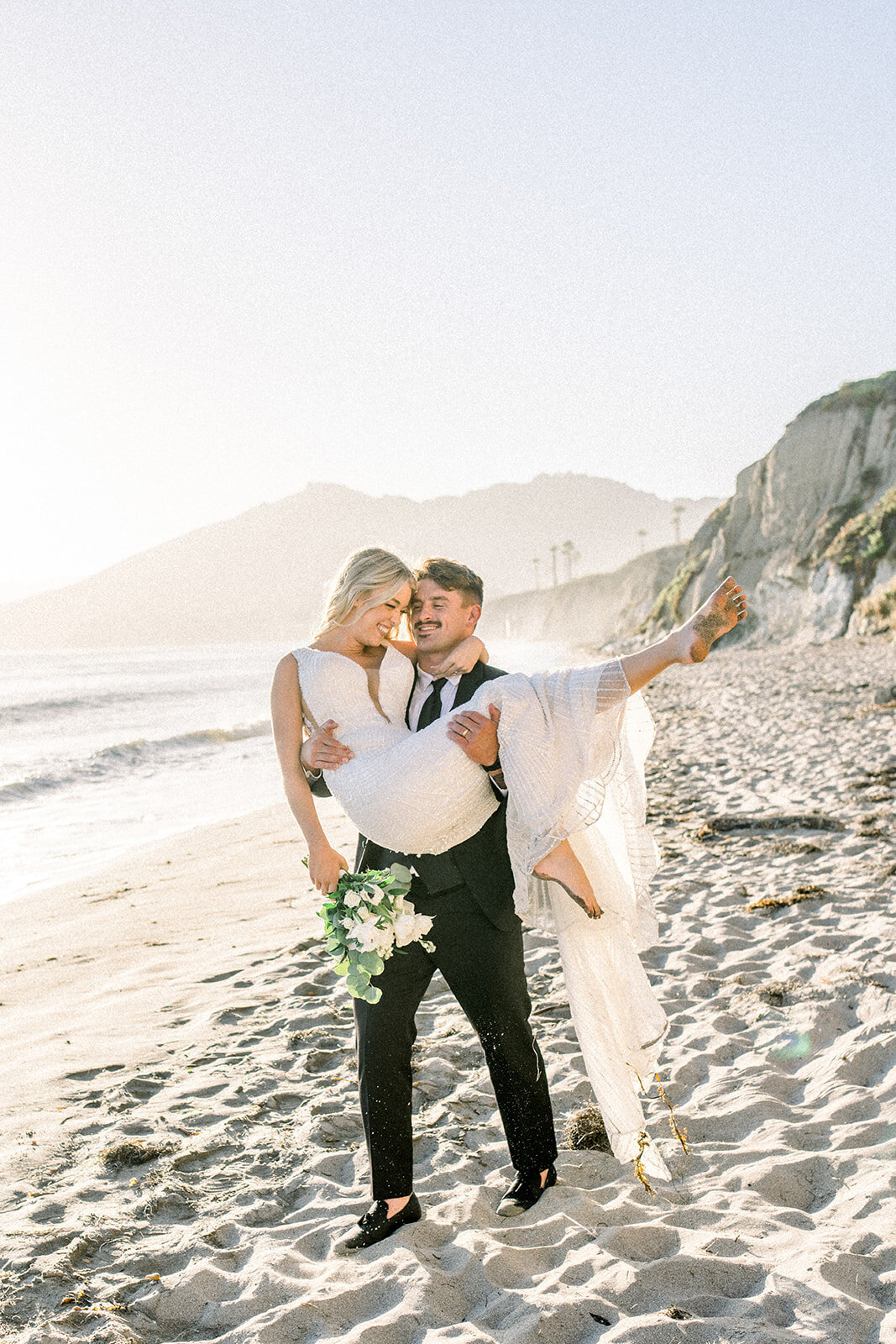 Groom carrying bride down the beach at Dolphin Bay Resort in Pismo Beach, CA