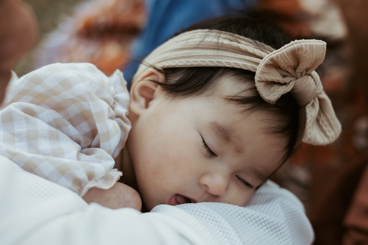 Sleeping four month old wearing a cute headband and fallen asleep in her dad's arms during their photography session.